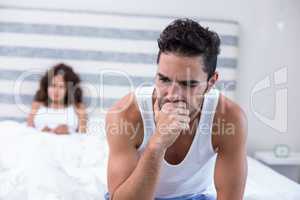 Tensed man sitting on bed while wife in background