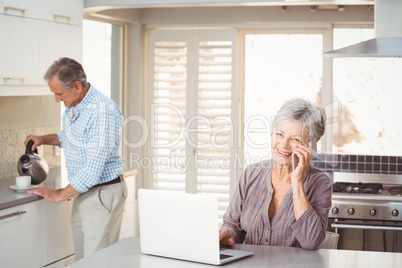 Senior woman talking on mobile phone with husband making tea in