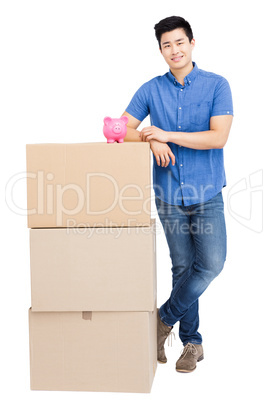 Young man standing near cardboard boxes with piggy bank