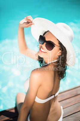 Beautiful woman wearing white bikini and hat sitting near pool