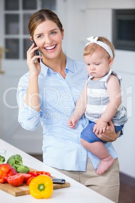 Woman with cellphone carrying daughter by kitchen counter