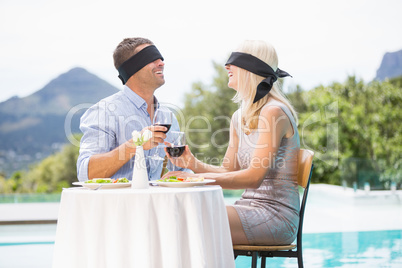 Blindfolded couple holding red wine while sitting by swimming po