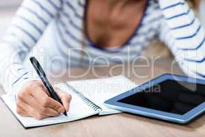 Midsection of woman writing in notebook by digital tablet