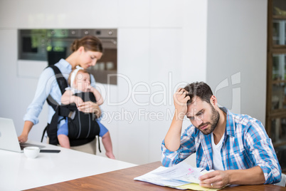 Frustrated man reading documents while wife and baby in backgrou