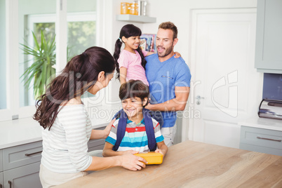 Mother giving lunch box to son with father carrying daughte