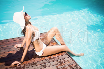 Beautiful woman wearing white bikini and hat sitting near pool