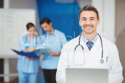 Portrait of smiling doctor with laptop and colleagues standing