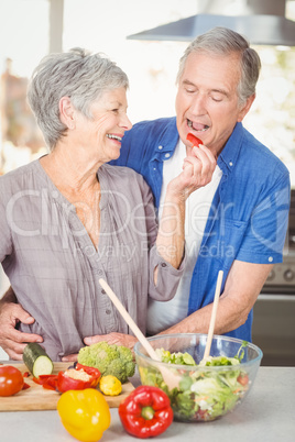 Happy senior woman feeding husband while standing at counter