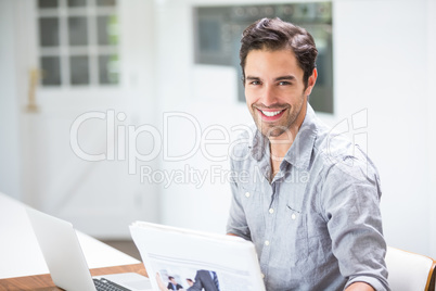 Smiling young man holding documents while sitting at desk with l