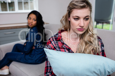 Upset woman holding pillow while female friend sitting on sofa