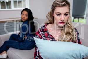 Upset woman holding pillow while female friend sitting on sofa