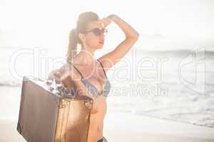 Glamorous woman with an old suitcase shielding eyes at beach