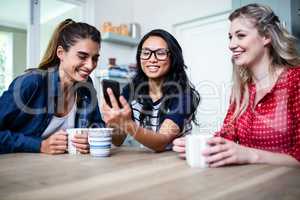 Cheerful female friends using mobile phone by table at home