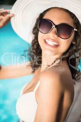 Beautiful woman wearing white bikini and hat sitting near pool