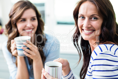 Cheerful female friends holding coffee mugs while sitting at tab