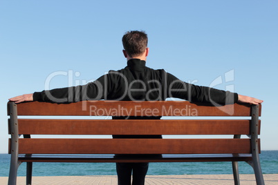 Mann sitzt in entspannter Haltung auf einer Bank am Meer