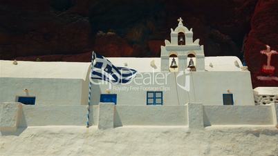 Greek Flag Flying Next to a Traditional White Cycladic Church with a Volcanic Red Cliff in the Background