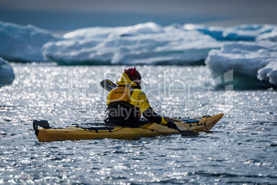 Backlit kayaker paddling past icebergs in sunshine