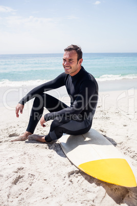 Happy surfer in wetsuit sitting with surfboard on the beach