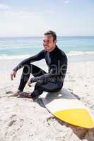 Happy surfer in wetsuit sitting with surfboard on the beach