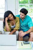 Upset couple sitting on sofa with laptop and bills