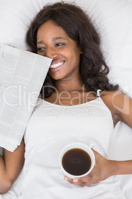 Beautiful young woman lying on bed holding coffee mug and news p