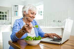 Senior woman eating salad while using laptop at table