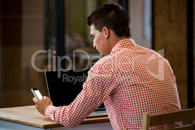 Man text messaging on mobile phone with laptop on desk