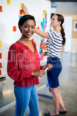 Young woman smiling at camera while colleague writing on white b