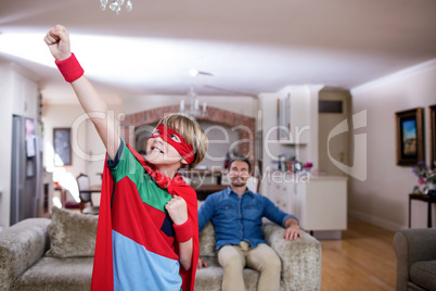 Son pretending to be a superhero while father sitting on sofa