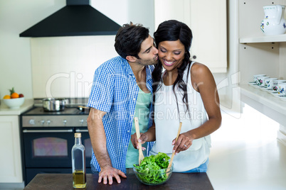 Man kissing a woman while mixing a salad in kitchen