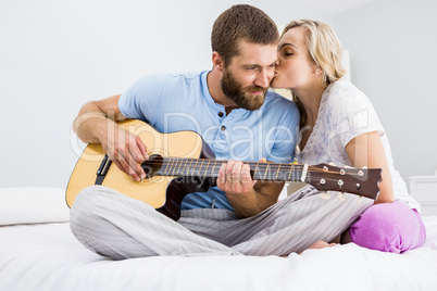 Man playing guitar while woman kissing him on cheek on bed