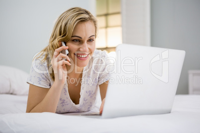 Woman using laptop while talking on mobile phone in bed