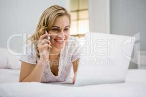 Woman using laptop while talking on mobile phone in bed