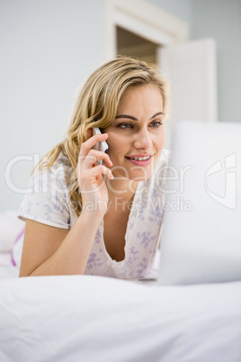 Woman using laptop while talking on mobile phone in bed