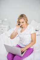 Woman using laptop while talking on mobile phone in bed