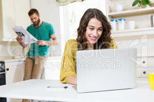 Woman using laptop while man reading newspaper in background
