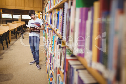 Young student reading book while standing near bookshelf