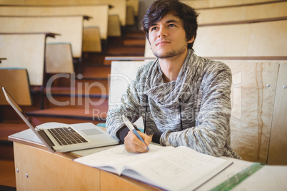 Thoughtful young student sitting at desk with book and laptop