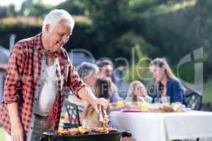 Senior man at barbecue grill while family having lunch in backgr