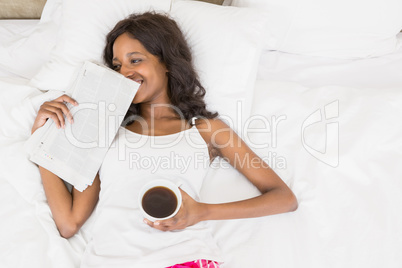 Beautiful young woman lying on bed holding coffee mug and news paper