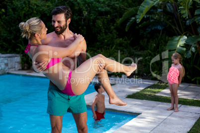 Man carrying woman at poolside with children in background