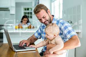 Father looking at baby boy while using laptop at home