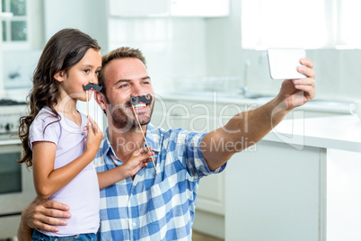 Father taking selfie with daughter holding artificial mustache