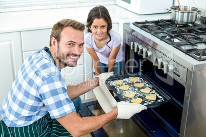 Father placing cookies tray in oven while daughter standing by h