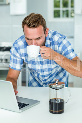 Man using laptop while drinking coffee in kitchen