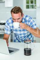 Man using laptop while drinking coffee in kitchen