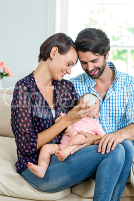 Parents feeding milk to baby boy while sitting on sofa