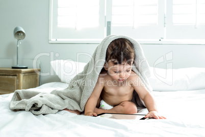 Baby boy covered with towel playing with digital tablet on bed