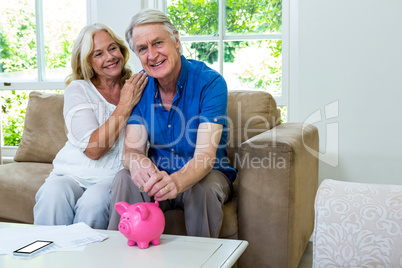 Smiling senior couple putting coin in piggi bank at home